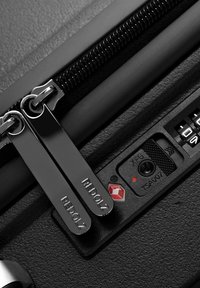 Primer plano de los tiradores de cremallera de una maleta negra etiquetados "REDOLZ" asegurados con un candado de combinación aprobado por la TSA con un logo de diamante rojo.