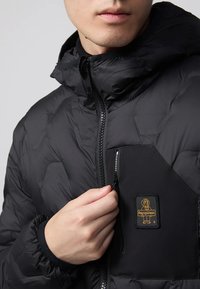 Giacca a vento nera con design trapuntato, chiusura frontale con zip e polsini elastici. Presenta un badge logo sulla tasca del petto.