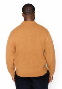 Strickpullover in einem warmen Karamellton mit lässiger Passform, hoher Kragen und geripptem Textur. Kombiniert mit dunklen Hosen.