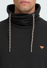 Hombre vestido con un hoodie negro de cuello alto, con cordones decorativos y un pequeño parche triangular marrón en el pecho.