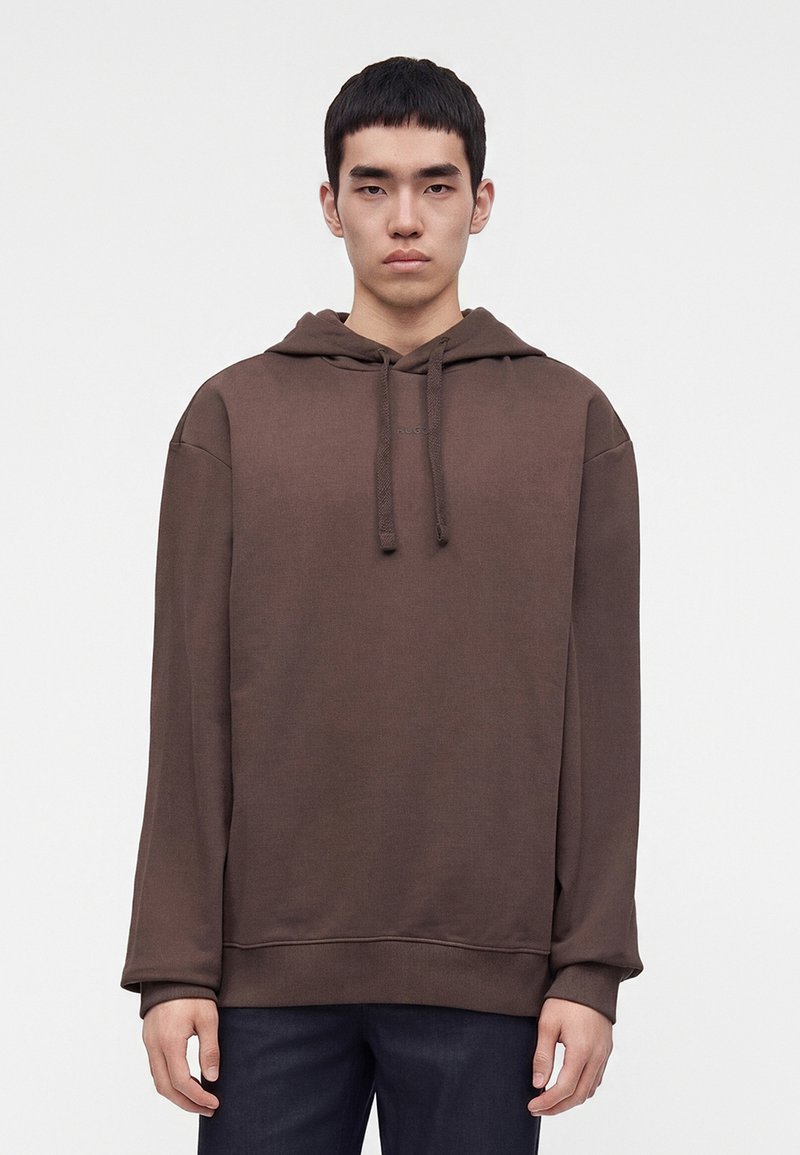 Jeune homme de face portant un sweat à capuche marron uni et un pantalon foncé devant un fond clair uni.