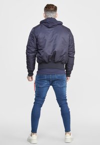 Chaqueta bomber azul marino con capucha, puños y dobladillo acanalados, con detalles acolchados en los hombros y cierre frontal con cremallera, combinada con vaqueros de corte slim.