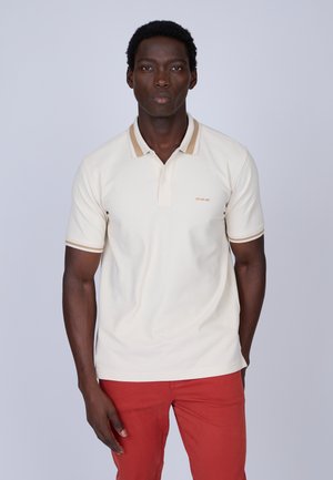 Polo beige avec col et poignets à rayures brunes, doté d'une patte de boutonnage à trois boutons et d'un petit logo sur la poitrine. Porté avec un pantalon rouge.