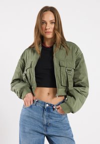 Zelena skraćena bomber jakna s dva prednja džepa, srebrnim detaljima i rebrastim manžetama, nosi se preko crnog topa i u paru sa plavim farmerkama.
