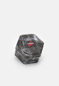 Conteneur hexagonal en étain avec une finition texturée noire et grise, présentant un logo G-SHOCK rouge et blanc sur le dessus et le côté.