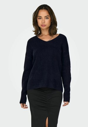 Kvinde iført en mørk navy V-hals sweater og et sort rynket skørt med frontslids, stående mod en ensfarvet lys baggrund.