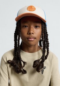 Gorra de camionero con un logo naranja sobre lona crema y lados de malla azul. La camiseta es beige con un logo triangular negro en el pecho.