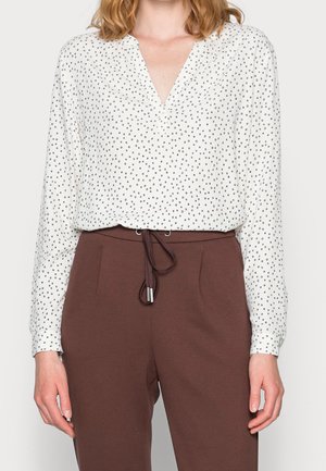 White long-sleeve blouse with small black polka dots tucked into high-waisted brown pants with a drawstring waist.