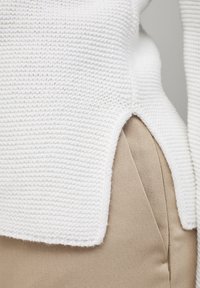 White knitted sweater featuring textured fabric with a side slit, paired with tan tapered pants, showcasing smooth material.
