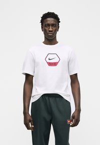 Νεαρός άνδρας που φοράει λευκό T-shirt Nike με κόκκινο λογότυπο εξαγώνου και σκουρόχρωμο αθλητικό παντελόνι με μικρό ταμπελάκι Nike σε λευκό φόντο.