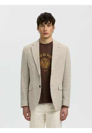 Mladý muž nosiaci béžové sako so vzorom houndstooth, hnedé tričko s grafikou a krémové nohavice, stojí pred jednoduchým svetlosivým pozadím.