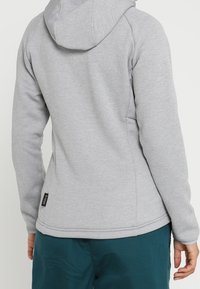 Ljusgrå huvtröja med en figurnära design, med ribbade ärmslut och en etikett på nederkanten till vänster. Mjuk material med en slät yta.