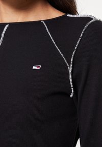 Svart räfflad långärmad topp med silvertrådsdetaljer. Har en liten broderad logotyp på vänster sida nära halsringningen.