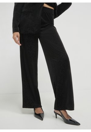Frau trägt schwarze, strukturierte, weit geschnittene Hosen und schwarze, spitze Slingback-Pumps aus Lackleder, steht mit einem Bein leicht nach vorne.