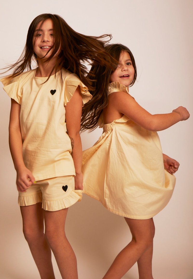 Helles gelbes Baumwolloutfit mit Rüschensäumen, Herz-Akzenten und lockerer Passform. Enthält ein Kleid und passende Shorts.