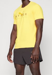 Camiseta atlética amarilla con cuello redondo y logo impreso "HYROX", combinada con pantalones cortos negros. Hecha de material ligero y que absorbe la humedad.