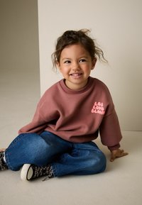 Ružová mikina s textom "Nech láska rozkvitne", voľný strih a rebrovanými manžetami. Kombinovaná s modrými džínsami a čiernymi vzorovanými topánkami, sediaca na neutrálnej podlahe.