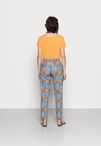 En orange kortärmad skjorta kombinerad med mönstrade byxor med blå och orange cirkulära mönster. Modellen bär platta sandaler.