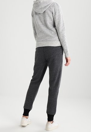 Osoba w szarej bluzie z kapturem i ciemnoszarych dresach z czarnymi mankietami, stojąca tyłem, ubrana w białe trampki na jednolitym tle.