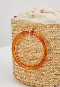 Sac rond en paille tissée avec dessus en tissu beige et grande anse circulaire translucide couleur ambre attachée par un anneau en or.