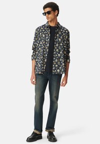 Camicia a maniche corte navy con fiori bianchi e gialli, abbinata a jeans denim scuri e scarpe nere. Il modello indossa occhiali da sole.