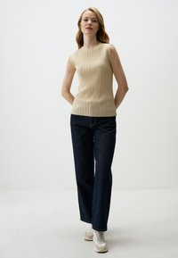 Top beige senza maniche con design a righe verticali, abbinato a jeans dritti blu scuro e sneakers bianche. Look semplice e moderno.