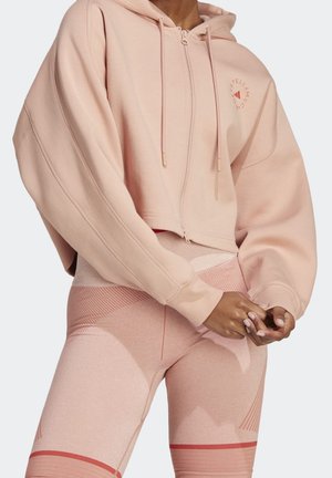 Personne portant un sweat à capuche court rose clair et des leggings taille haute assortis avec des détails subtils à motifs, mains jointes devant.