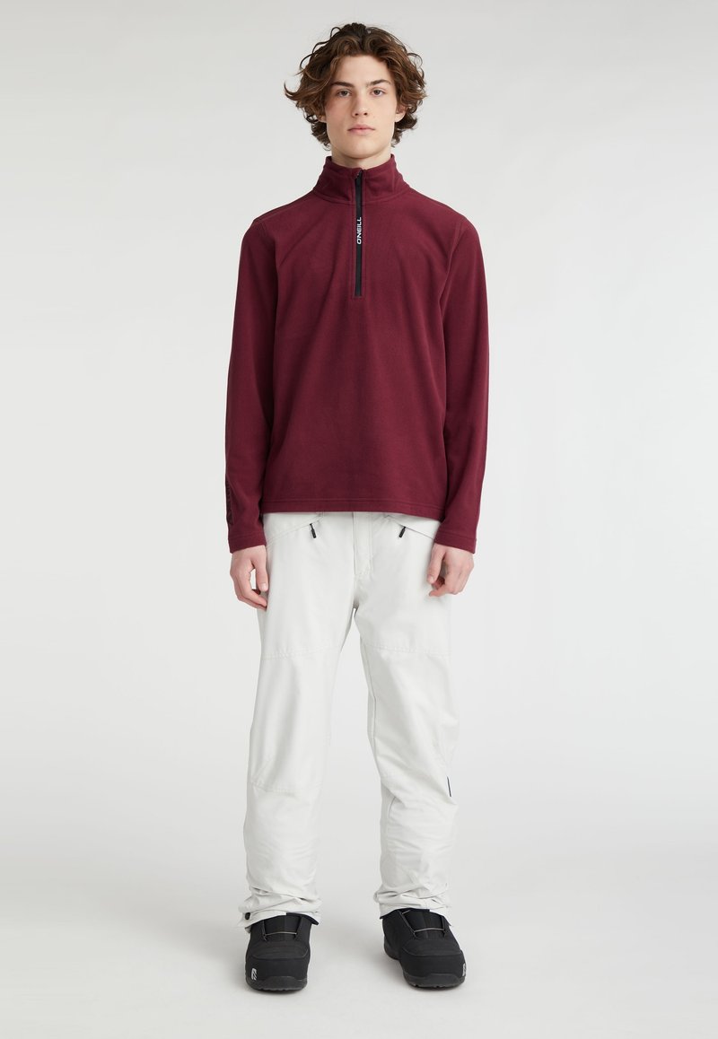 Pull en polaire bourgogne avec col zippé, assorti à un pantalon blanc et des bottes noires. Texture douce, design simple et détails fonctionnels.