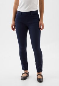 Tamsiai mėlyni siaurėjančio silueto kelnės su lygiu audiniu, turinčios juosmenį ir šonines kišenes, derinamos su juodais bateliais