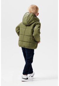 Olivengrøn pufferjakke med hætte, quiltet design og refleksdetaljer, parret med mørkeblå jeans og hvide sneakers.