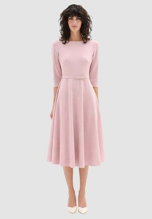 Donna con capelli ricci e scuri che indossa un vestito rosa pallido fino al ginocchio con maniche a tre quarti, cintura e tacchi bianchi a punta, in piedi su uno sfondo grigio.