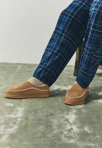 Pruun suede libisemisjalanõud kreemjate õmblustega ja struktureeritud tallaga, kantud beežide sokkide ja sinise ruudulise püksiga pehmest hallist pinnast.