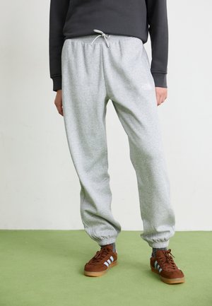 Lichtgrijze joggingsbroek met elastische boorden, een trekkoord in de taille en logo-detail. Gecombineerd met bruine sneakers met blauwe strepen.