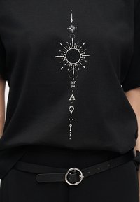 Μαύρο πουκάμισο με λευκά ουράνια και αστρολογικά σύμβολα τυπωμένα κάθετα, σε συνδυασμό με μαύρο παντελόνι και μαύρη ζώνη με ασημένια στρογγυλή αγκράφα.