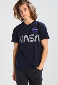 Camiseta de algodón azul marino con un gran logo gris de "NASA" y un parche circular de NASA en el pecho. Manga corta y cuello redondo.