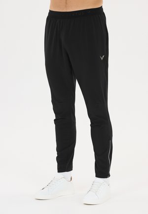 Mann trägt schwarze Jogginghose mit kleinem Logo am Oberschenkel und weiße Sneaker, steht vor schlichtem weißem Hintergrund.