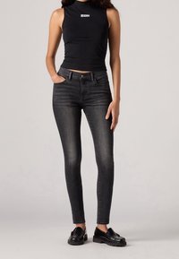 Černý top bez rukávů s bílým logem, v kombinaci s tmavě šedými úzkými džínami. Džínov�é kalhoty mají klasický design s pěti kapsami a mírně vybledlý vzhled.