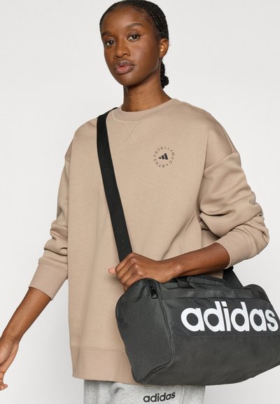 Μαύρη τσάντα Adidas τύπου duffel με λευκό λογότυπο. Άτομο φορώντας μπεζ φούτερ με κυκλικό λογότυπο, μανίκια σηκωμένα, κρατώντας την τσάντα με το ένα χέρι.