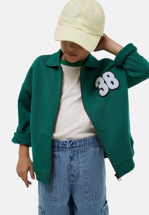 Enfant portant une casquette jaune clair, une veste zippée verte avec un écusson "38", une chemise blanche et un pantalon en jean bleu, la main levée vers la tête.