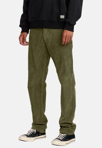 Olive green corduroy pants with straight legs, soft texture, and front pockets. Worn with a black sweatshirt and black sneakers.