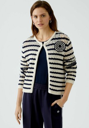Gestreifter Cardigan in Creme und Marineblau, mit floralem Häkelakzent an der Schulter, gestrickter Textur und einem knopflosen Frontverschluss.