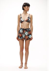 Γυναίκα που στέκεται ξυπόλητη, φοράει μαγιό μπικίνι τοπ και σορτς με ταιριαστό σχέδιο σε χρώματα μαύρο, καφέ, μπλε και άσπρο, κοιτάζοντας προς τα πλάγια.
