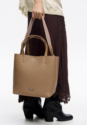 Bolso tote de cuero beige con asas beige tejidas, presenta una textura lisa, forma estructurada y logo plateado en el frente.