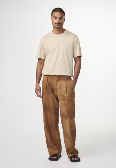 T-shirt beige a maniche corte, pantaloni a quadri marroni con una finitura testurizzata e sandali neri, indossati da un uomo che si trova davanti a uno sfondo semplice.