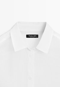 Bílá košile s límečkem a knoflíky, s černou štítkem s nápisem „Massimo Dutti“ a „Made in Cambodia“ uvnitř límce.