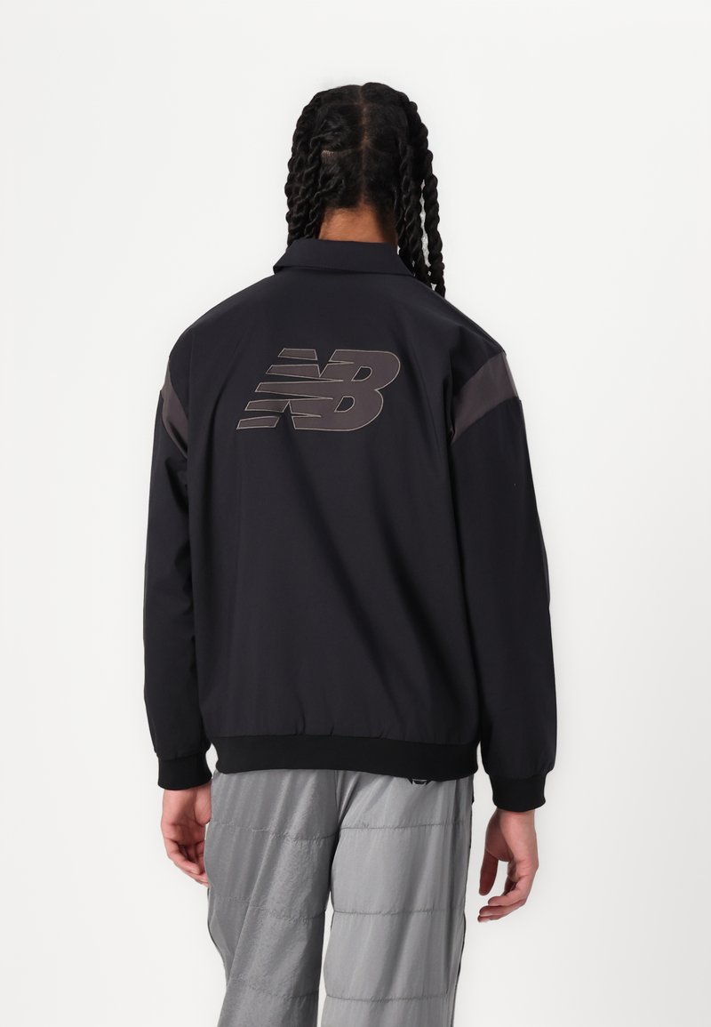 Chaqueta bomber negra con puños y dobladillo acanalados. Presenta un gran logo reflectante "NB" en la parte posterior y paneles de hombro en contraste.
