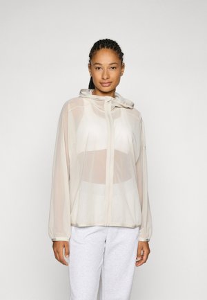 Lightweight, sheer beige hooded jacket with a full zipper, elastic cuffs, and a slightly loose fit, worn over gray sweatpants.