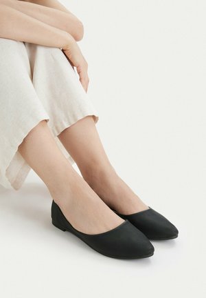 Füße in schwarzen spitzen Ballerinas, die Beine sind übereinandergeschlagen und tragen helle, weite Hosen, vor einem schlichten Hintergrund.