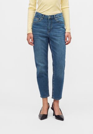 Person, die hoch taillierte blaue Jeans, ein blassgelbes Langarmshirt und schwarze Slingback-Pumps mit spitzer Kappe trägt, steht vor einem schlichten Hintergrund.