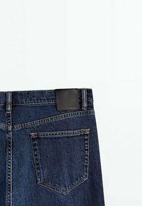 Jean denim bleu foncé montrant la poche arrière et la boucle de ceinture avec un patch de marque rectangulaire noir sur fond blanc.
